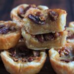 Butter tarts sitting on top of each other. The top ones are cut in half to show the insides. More tarts are scattered around in the background.