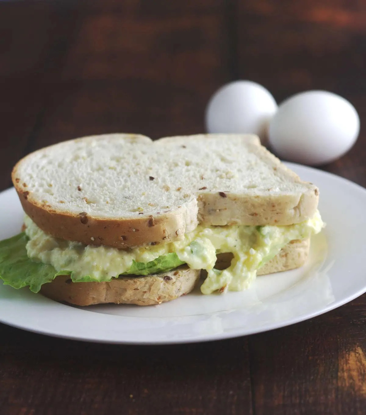 A intact sandwich on a white plate.  Two eggs are in the background.