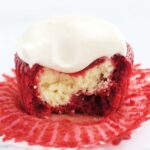 A cupcake sitting on it's opened cupcake liner. There is a bite out of the cupcake so you can see it's cheesecake filling.