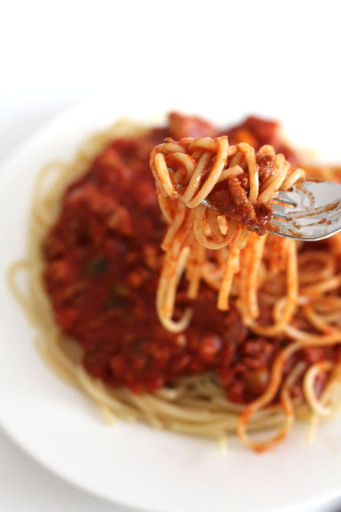 A forkful of spaghetti and meat sauce with a plate of it in the background.