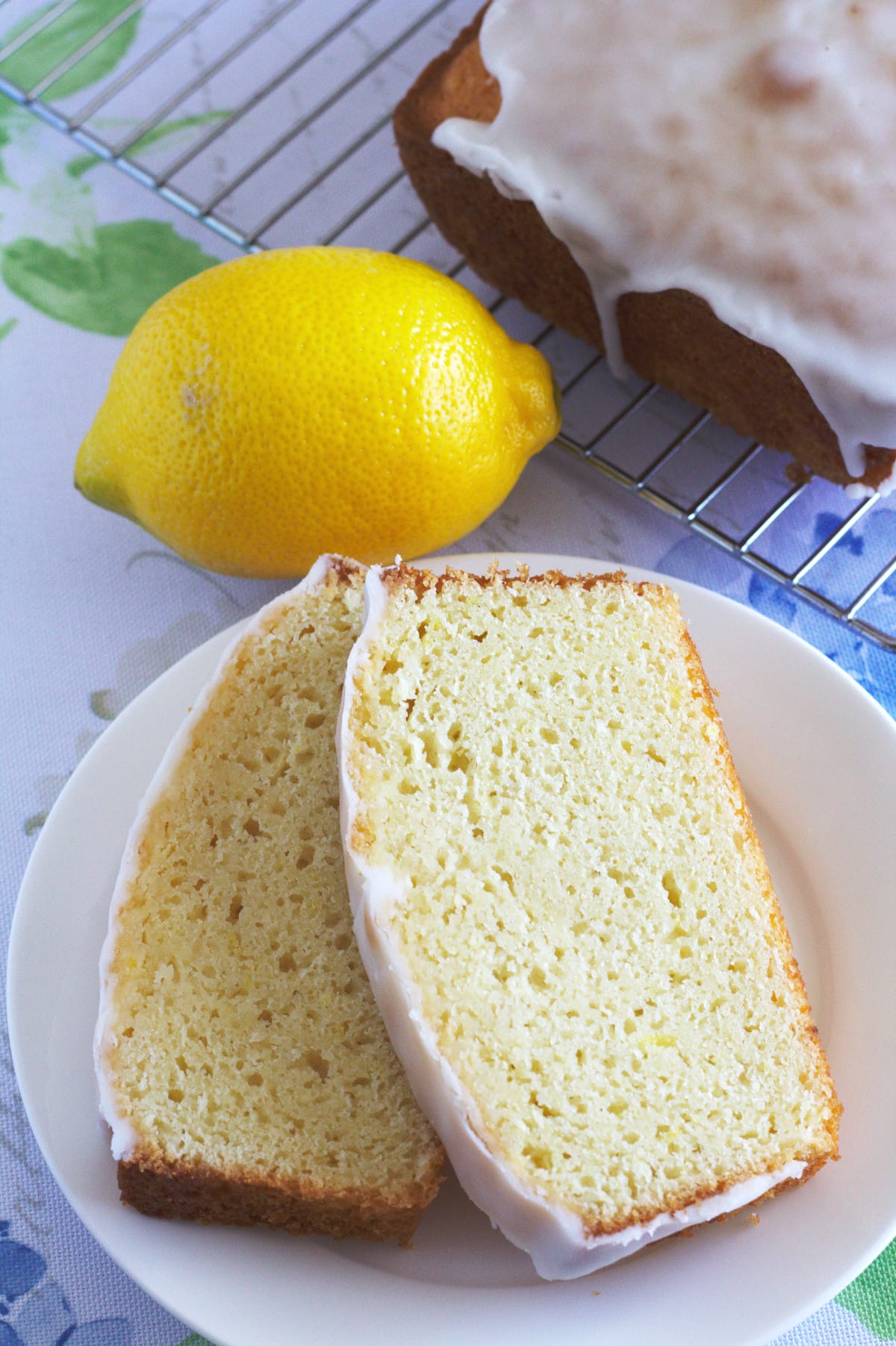 Two slices of lemon loaf on a plate with a lemon and the rest of the loaf.