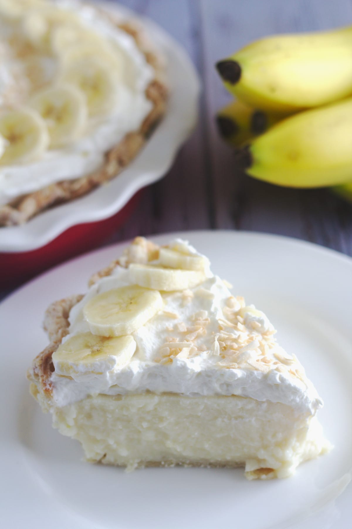 A slice of pie on a plate.  Bananas and the rest of the pie are in the background.