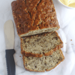 Banana bread with some slices cut from it. A spreading knife lays on one side of the bread and a bowl of butter on the other side.