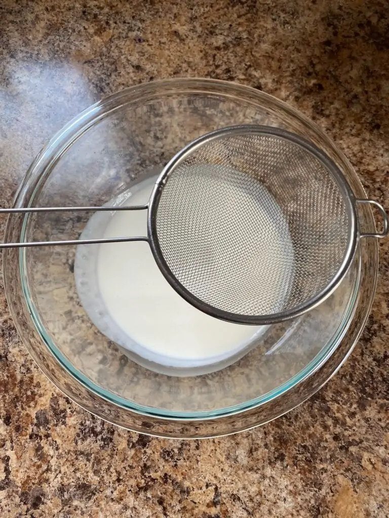 Heavy whipping cream in a medium clear bowl with a fine sieve over it.