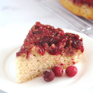 A slice of cake on a white plate with three whole cranberries in front of it.
