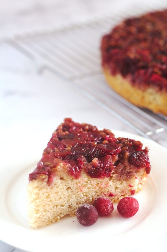 A slice of cake on a white plate with three whole cranberries in front of it.  The rest of the cake is seen partially in the background.
