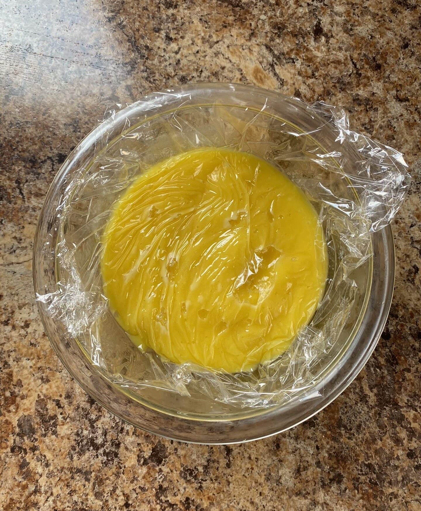 Lemon curd in a small clear bowl. Plastic wrap is placed directly on the lemon curd.