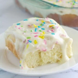 A cinnamon roll on a white plate. The cinnamon roll is frosted and has sprinkles on top.