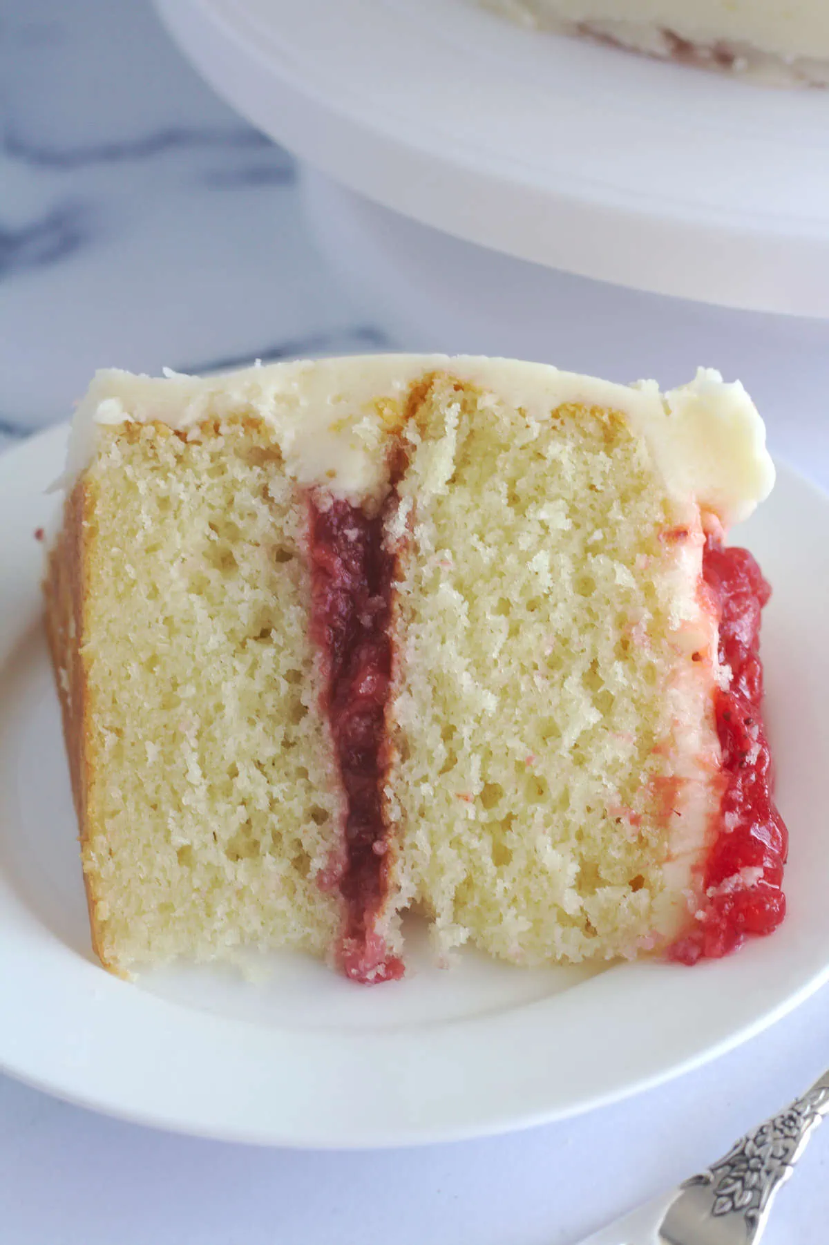A slice of cake on a white plate. The cake has strawberry filling in the middle of the cake, and on top.