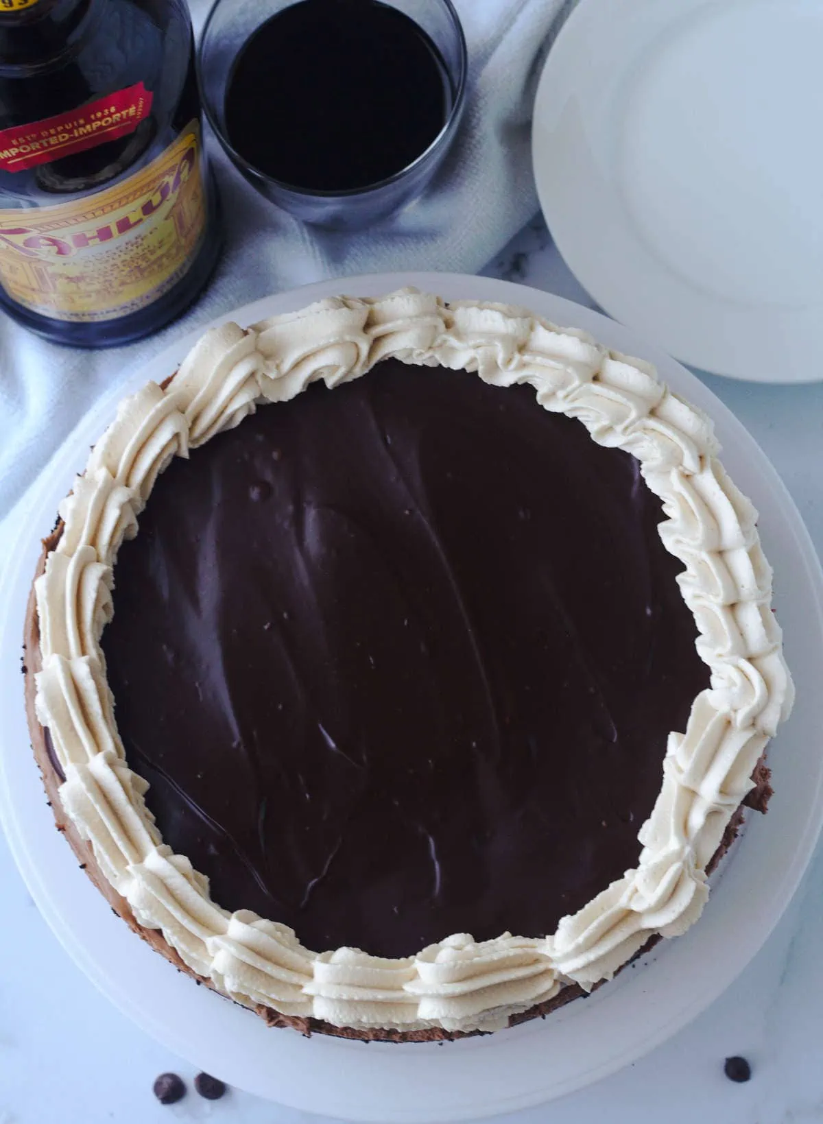 A top view of a cheesecake.  A Kahlua liquor bottle is to the top left of the cake.  Beside it is a glass filled with Kahlua.  A pile of dessert plates or on the top right of the cake.