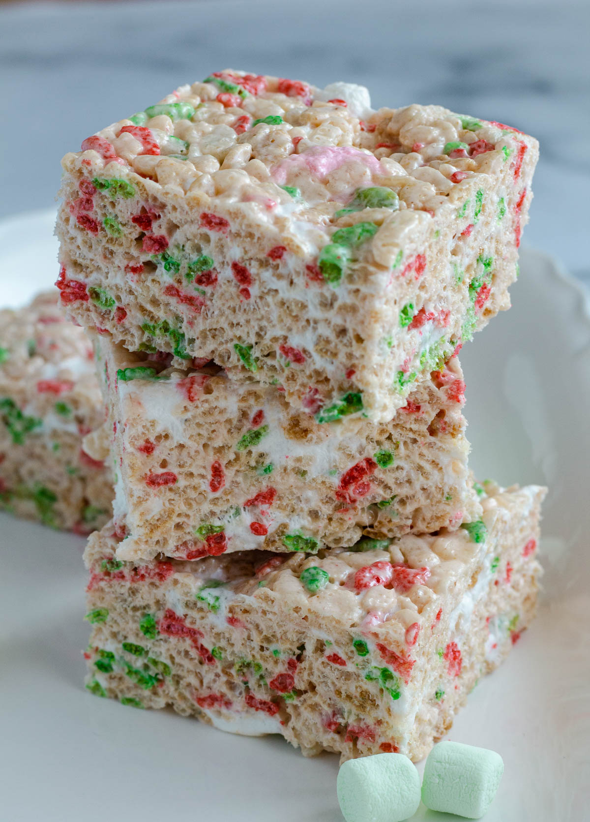 Three Rice Krispie squares sitting on top of each other on a white plate.  Two green marshmallows sit in front of them.