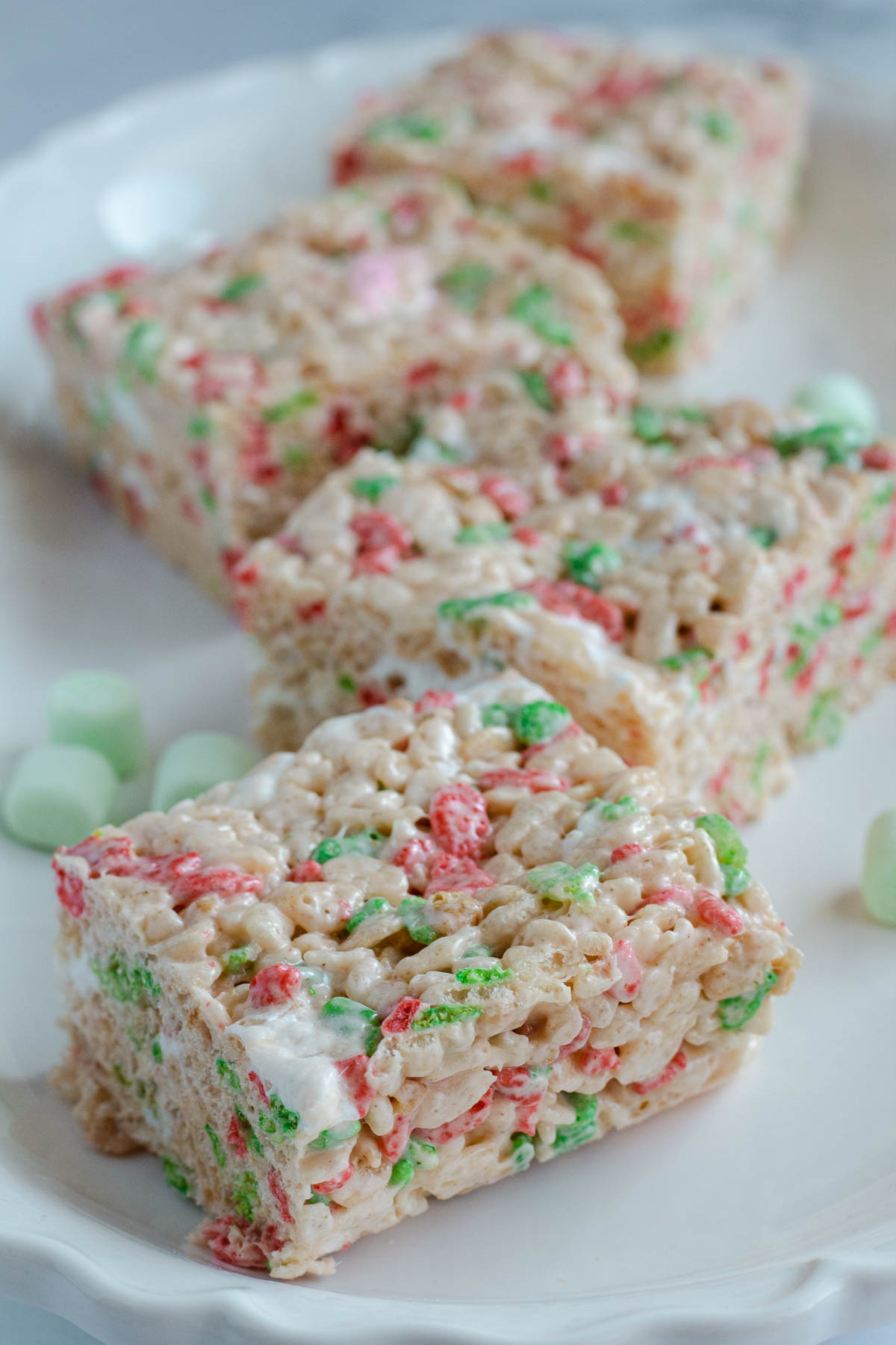 Four Rice Krispie squares laid out on a white plate. Green marshmallows are scattered amongst them.
