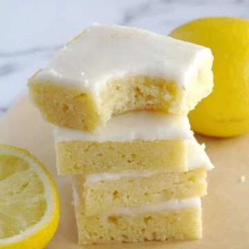 Four lemon brownie squares stacked on top of each other. The top brownie has a bite taken from it. Two lemons sit around the brownies.