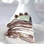 A slice of crepe cake on a white plate. The rest of the cake is in the background on a white cake stand.