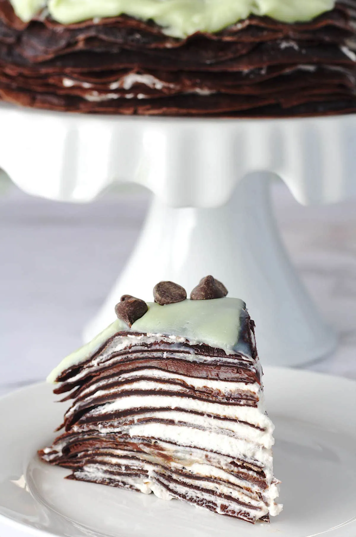 A slice of crepe cake on a white plate.  The rest of the cake is in the background on a white cake stand.