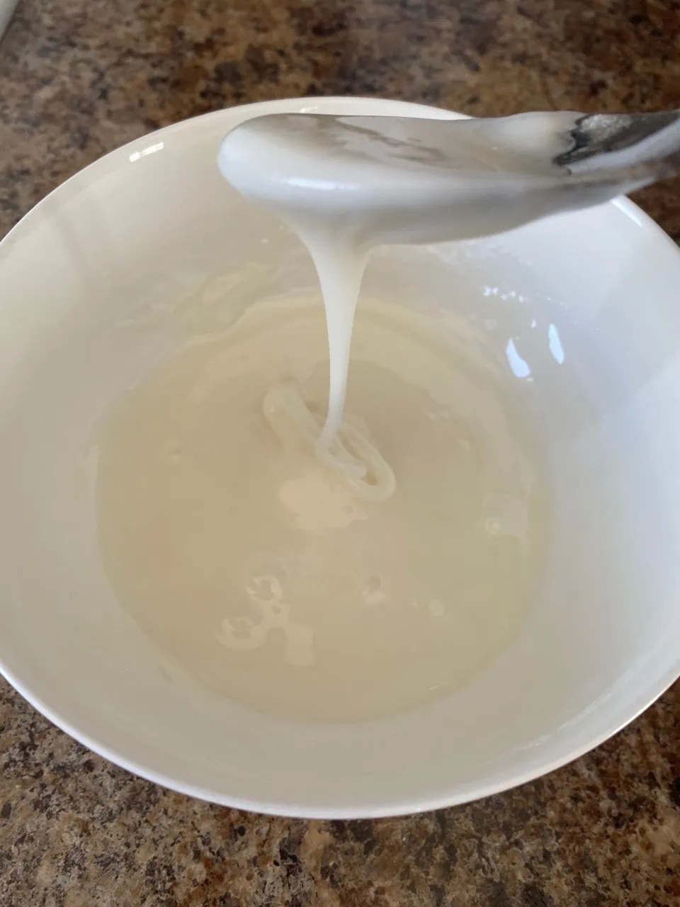 Glaze for brownies in a white bowl.  A spoon is above the bowl and glaze is running off the spoon, back in to the bowl.