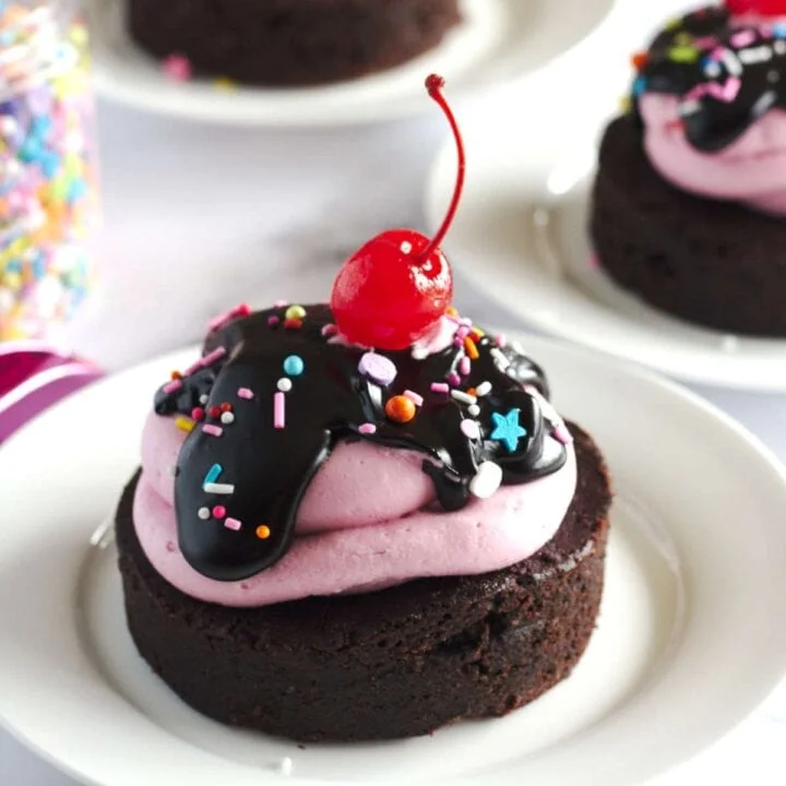 A mini brownie cake on a white plate. A stemmed cherry sits on top of the cake.