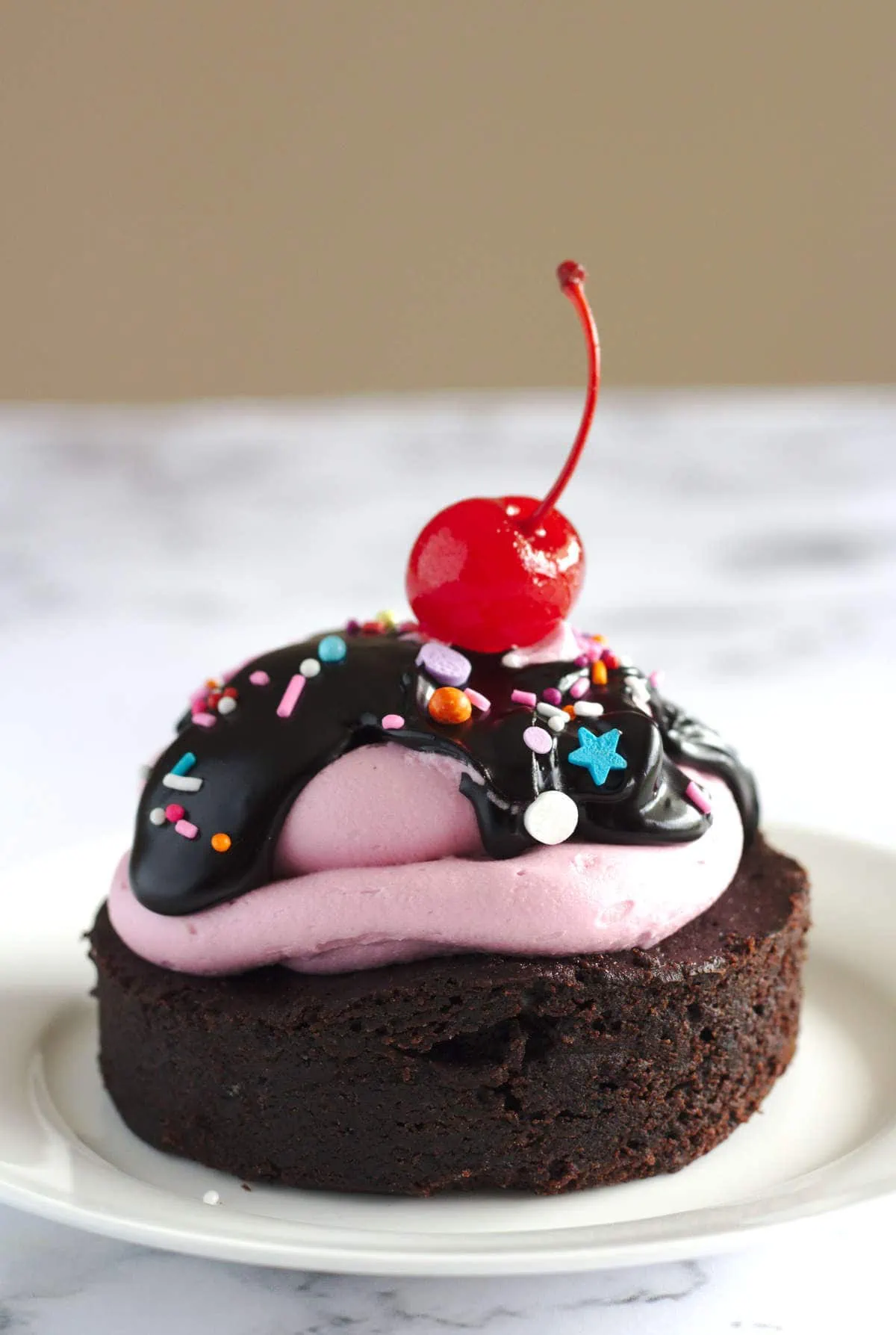 A mini brownie cake on a white plate. A stemmed cherry sits on top of the cake.