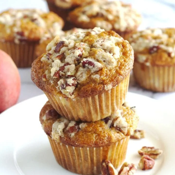 Two muffins with one sitting on top of the other. They sit on a white plate with some pecans on it.