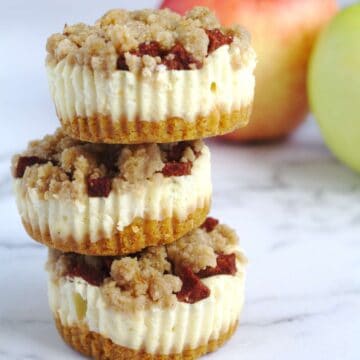 Three mini cheesecakes, on top of each other. Two apples sit in the background.