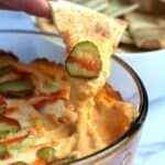 A dip with a person's hand dipping a pita chip into it.