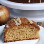 A slice of coffee cake on a white plate. The rest of the cake is in the background on a white cake plate.