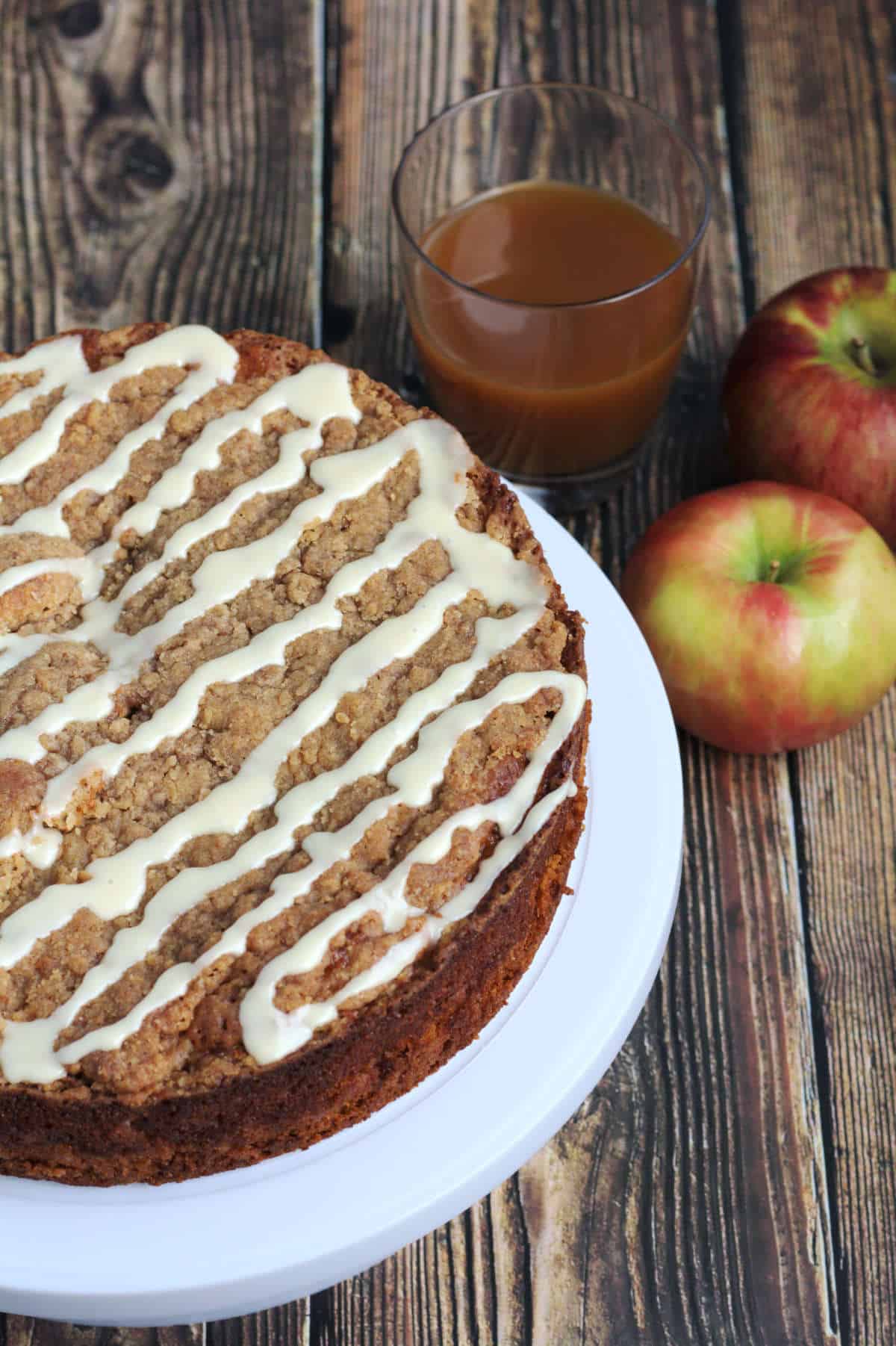 A partial view of a coffee cake on a white stand. A clear glass of apple cider and some apples sit beside it.
