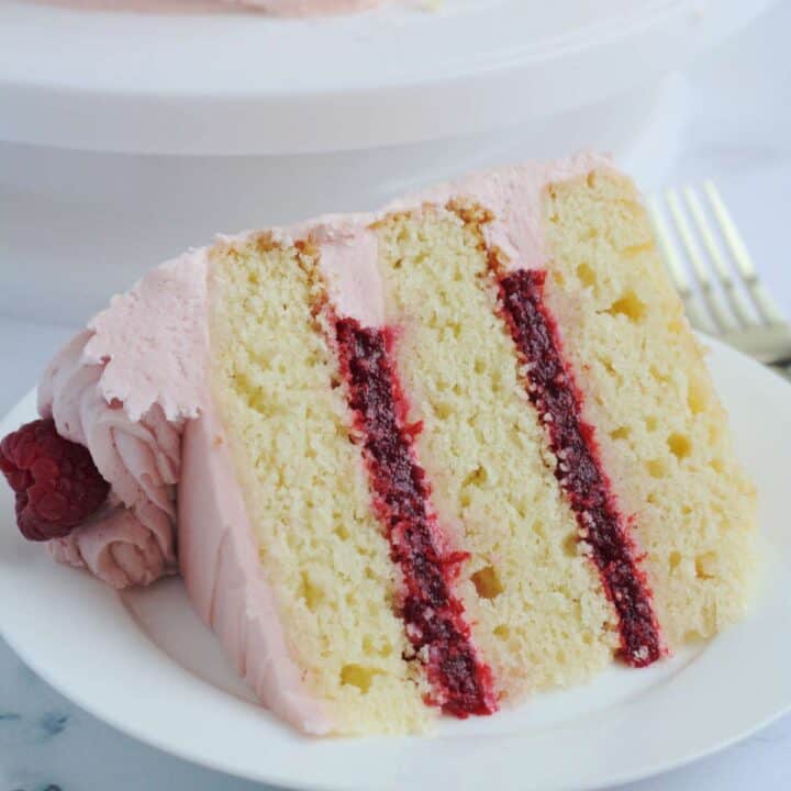 A slice of cake on a white plate. The rest of the cake is in the background on a cake table.