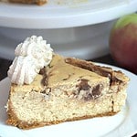 A slice of cheesecake on a white plate. The rest of the cheesecake is in the background on a white cake plate.