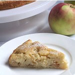 A slice of cake on a white plate. An apple and the rest of the cake are partially seen in the background.