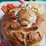 The bread bowl dip on a white platter with various vegetables and chips around it. A hand is holding a chip with some dip on it.