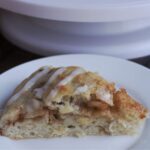 A slice of focaccia on a white plate. The rest is in the background on a white cake stand