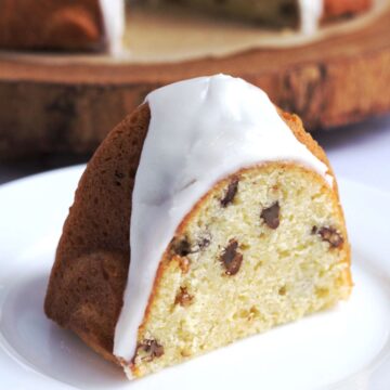 A slice of cake on a white plate. The rest of the cake is seen partially in the background.