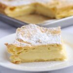 A vanilla custard square on a white plate. The rest of them are in a baking pan in the background.