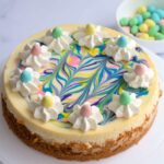 A full cheesecake on a white cake stand. Some mini eggs are in a white bowl in the background.