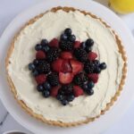A full tart on a white cake plate. Some lemons and a bowl of berries sit in the corners of this picture.