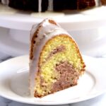 A slice of cake on a white plate. The rest of the cake sits in the background.