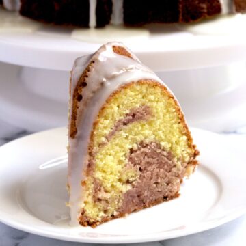 A slice of cake on a white plate. The rest of the cake sits in the background.