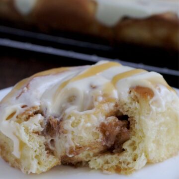 A cinnamon roll that has a bite out of it on a white plate. A clear baking pan with the rest of the cinnamon rolls in it, sits behind the plate.