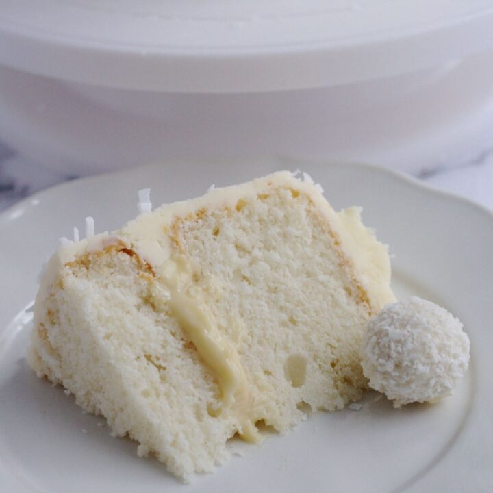 A slice of cake on a white plate. A raffaello truffle sits on the plate too.