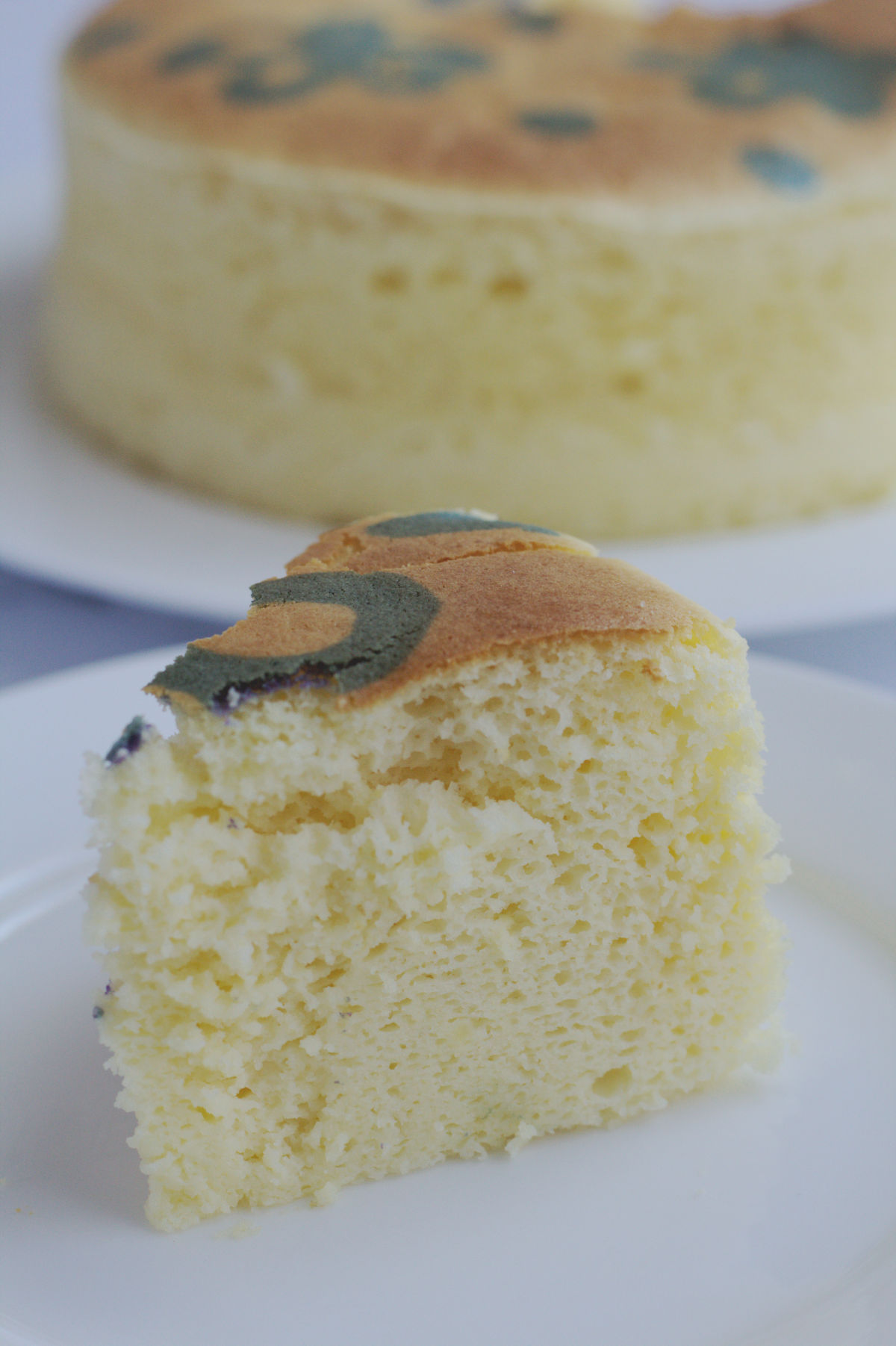 A slice of cheesecake on a white plate. The remainder of the cake is in the background on a white cake plate.