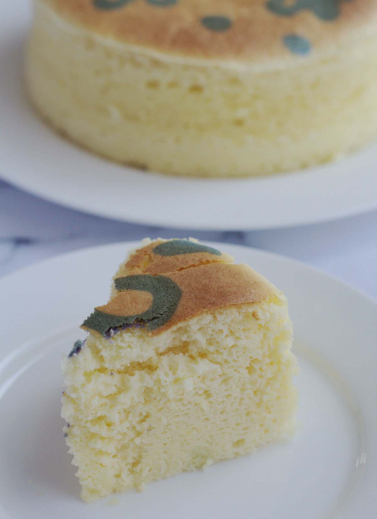A slice of cheesecake on a white plate.   The remainder of the cake is in the background on a white cake plate.