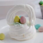 A meringue Easter basket on a white plate. Some mini eggs are around the basket.
