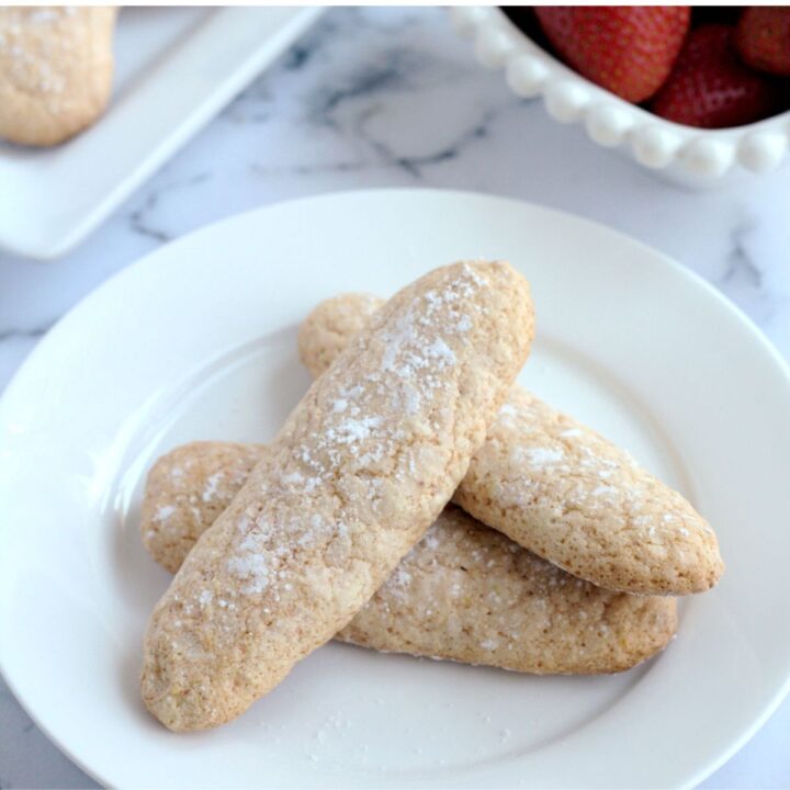 Ladyfinger cookies on a white plate. Some strawberries are in a white dish in the background.