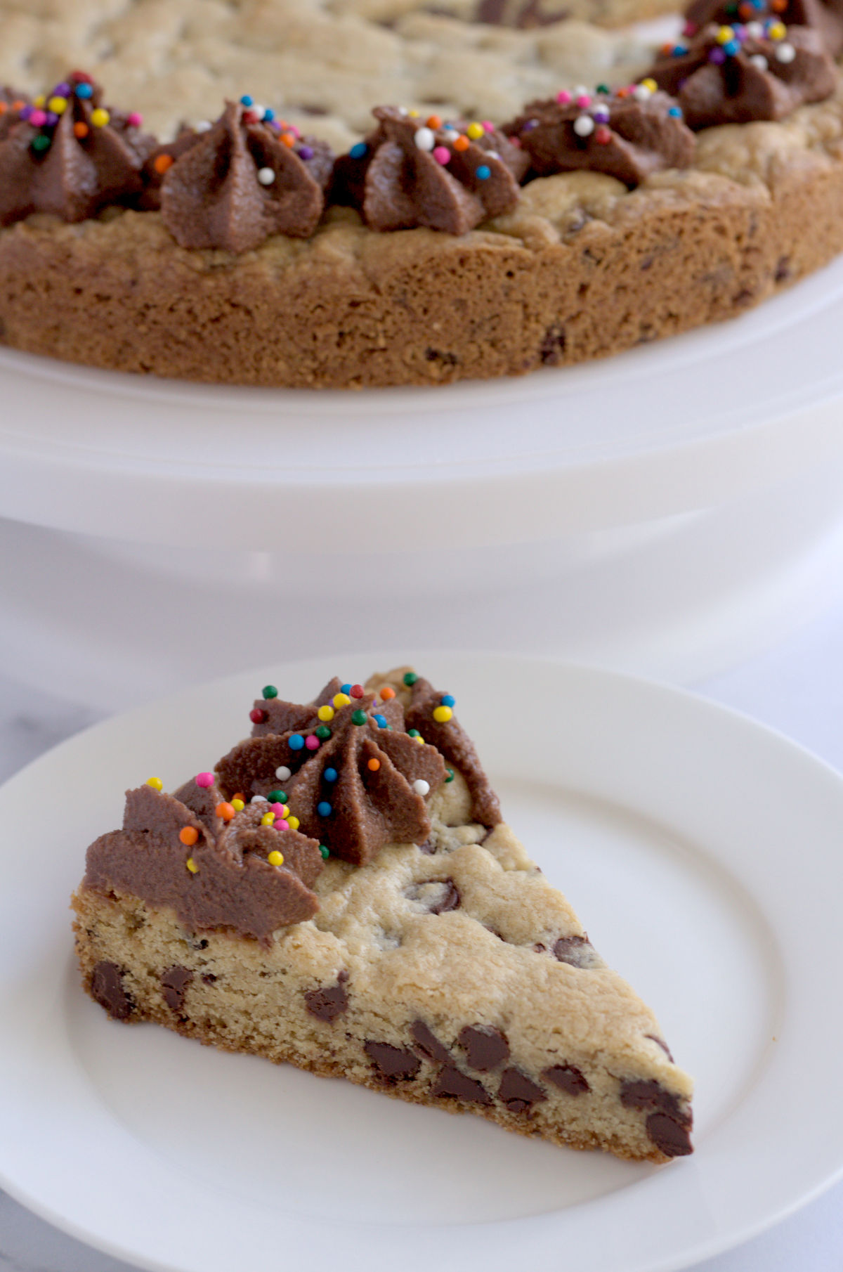 A slice of cookie cake on a white plate. The rest of the cake is seen partially in the background on a white cake plate.