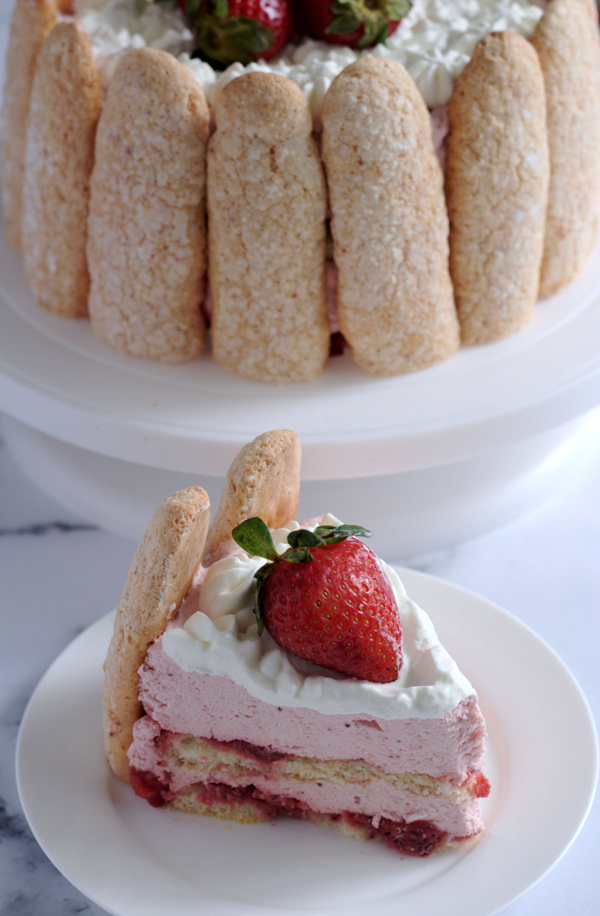 A slice of cake on a white plate. The rest of the cake is in the background on a white cake stand.