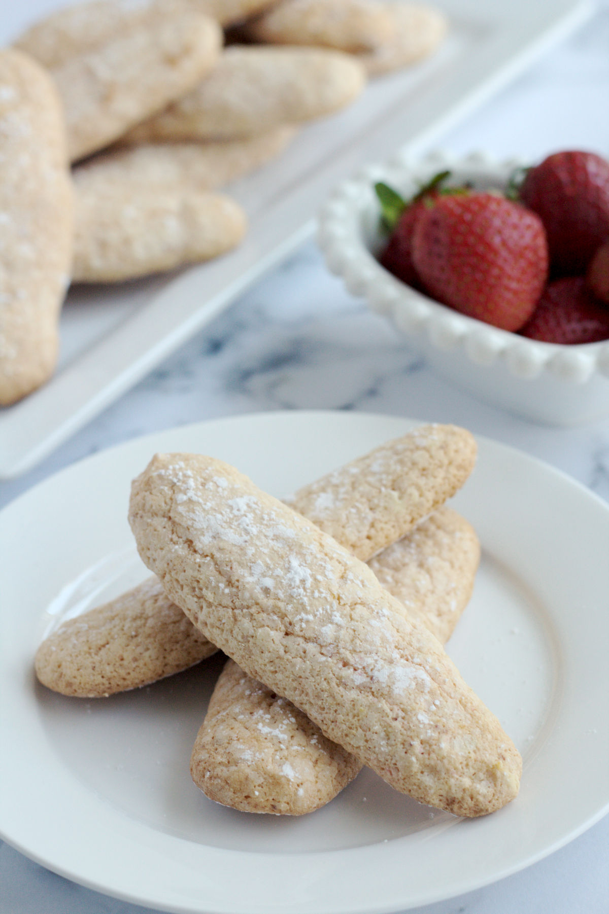 Ladyfinger cookies on a white plate. Some strawberries are in a white dish in the background.