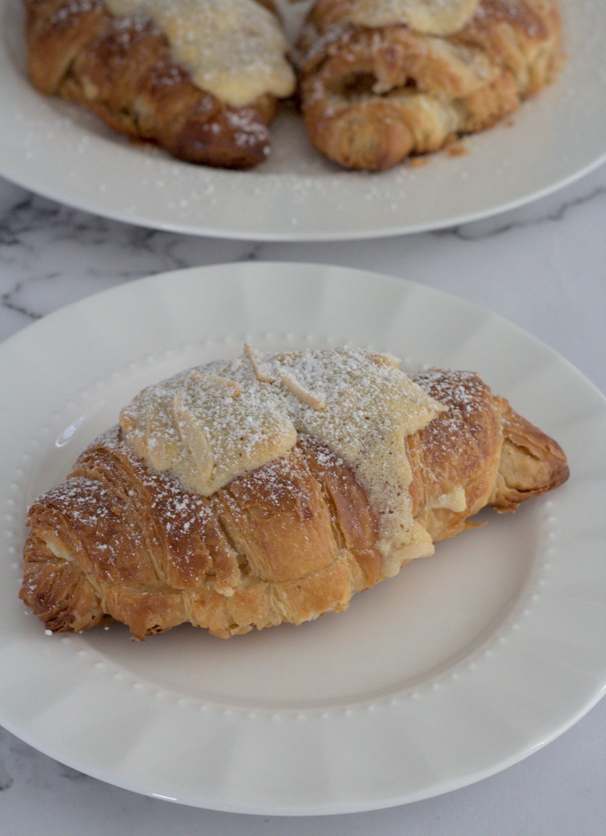 A single croissant on a white plate. Two more croissants are in the background on a large white plate.